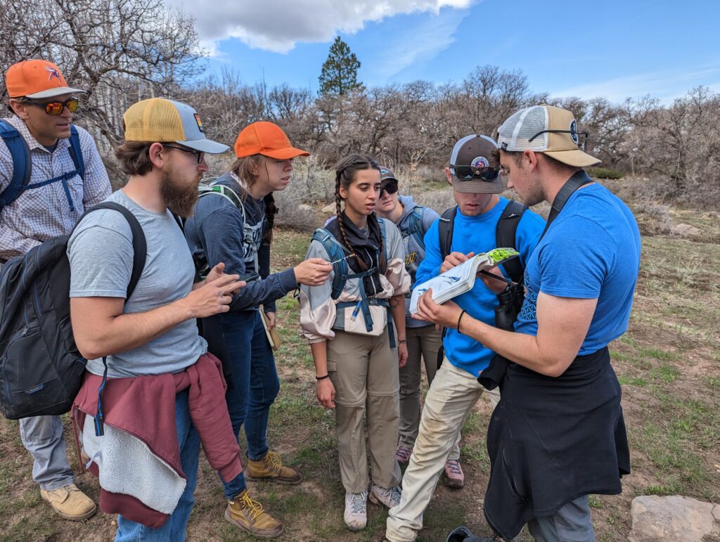 School of Science & Agriculture Students in ball caps gather around a classmate holding a field guide on a desert trail.